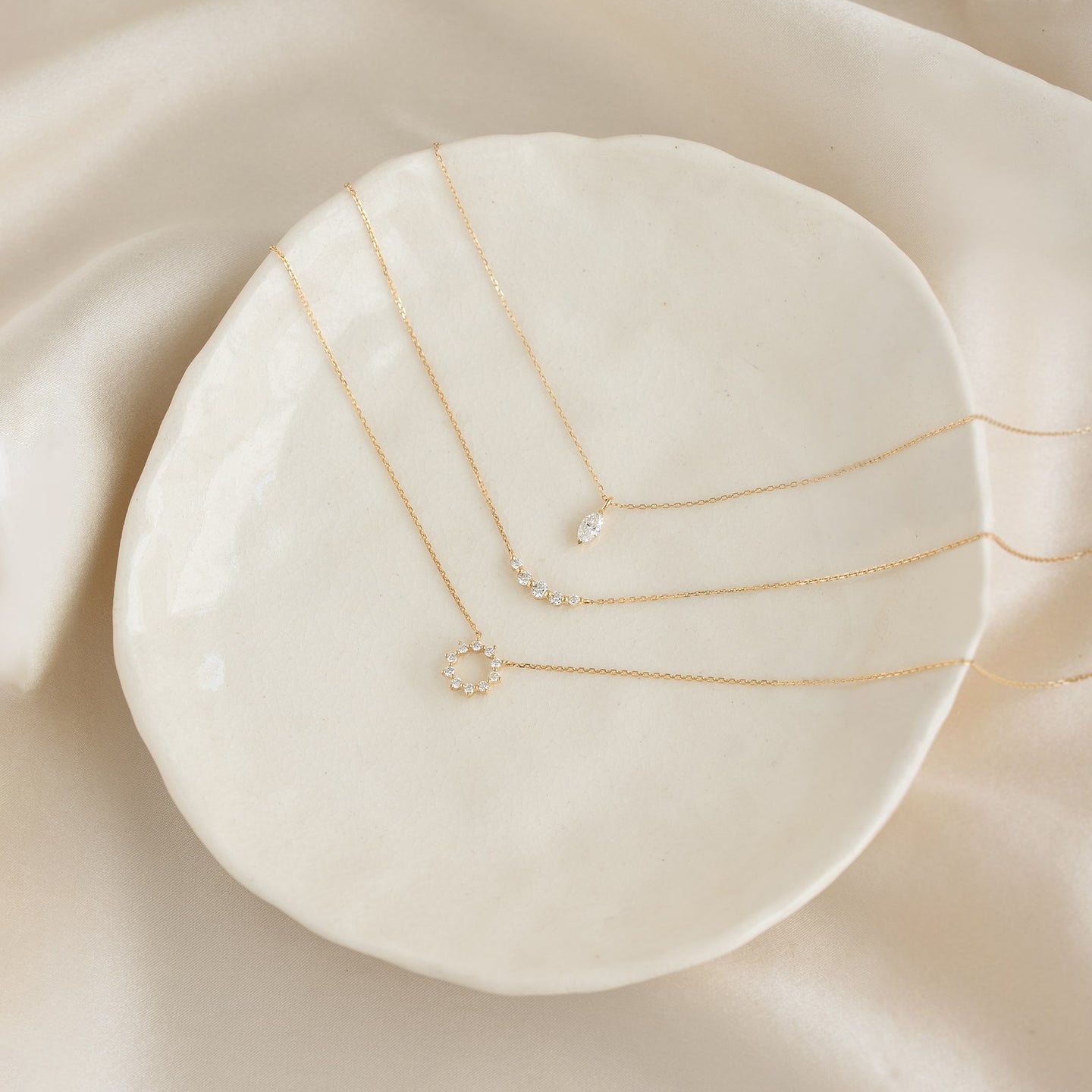 Close-up of three delicate gold necklaces including the Marquise Diamond Necklace 14k Gold - Violetta on a white ceramic dish