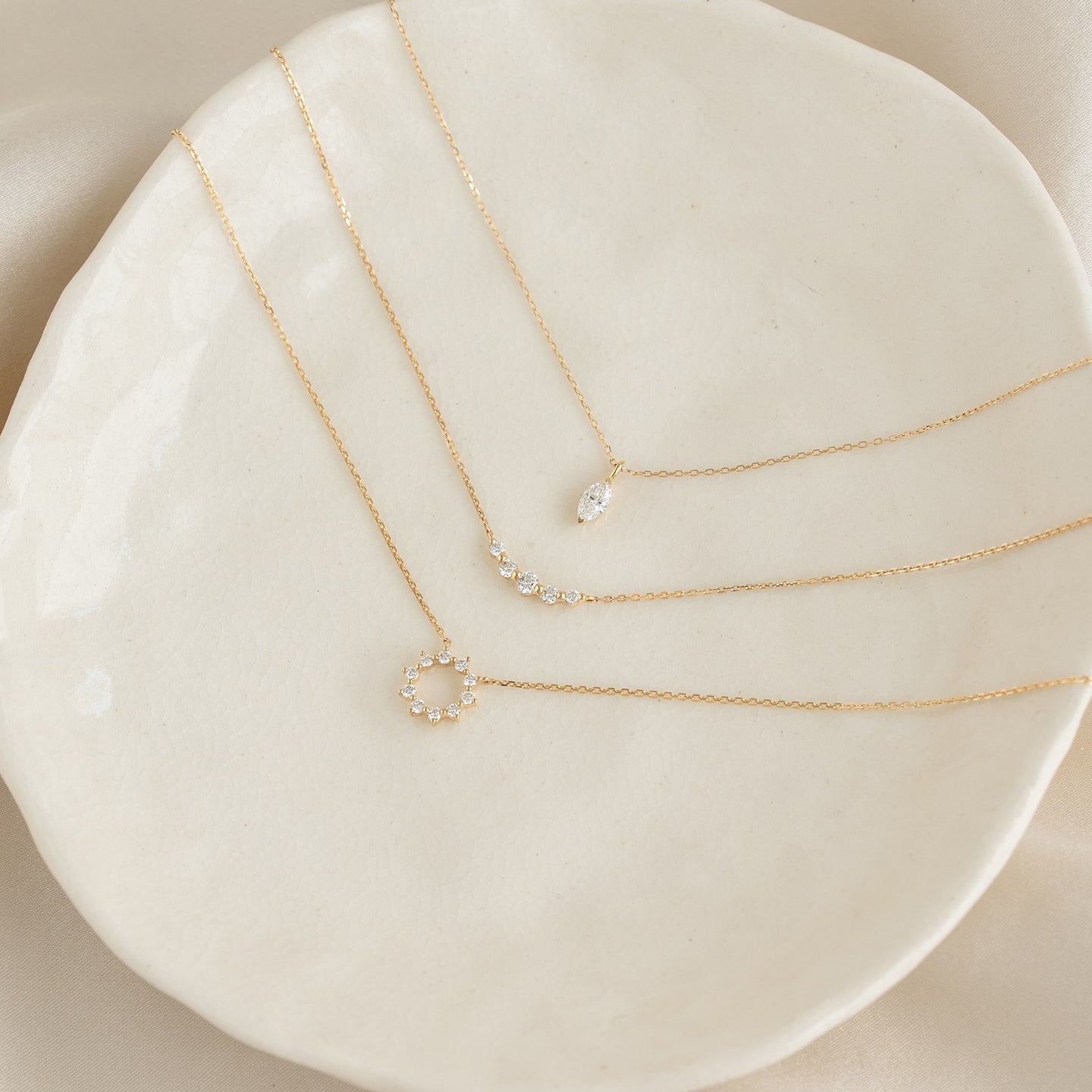 Close-up of three delicate gold necklaces with diamond pendants displayed on a cream ceramic plate, Diamond Sun Necklace 14k Gold - Leda