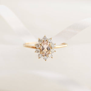 Close-up of a delicate gold ring with a central oval peach gemstone surrounded by small clear stones on a soft background