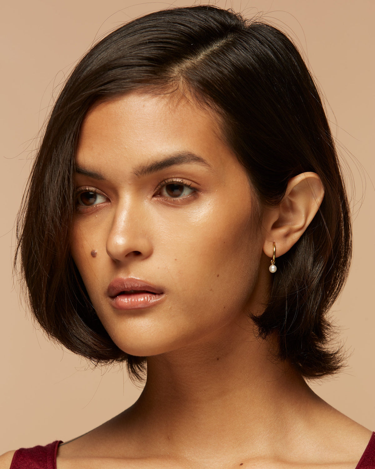 A person with short brown hair and pearl earring gazes thoughtfully to the side against a beige background. The expression is calm and introspective.