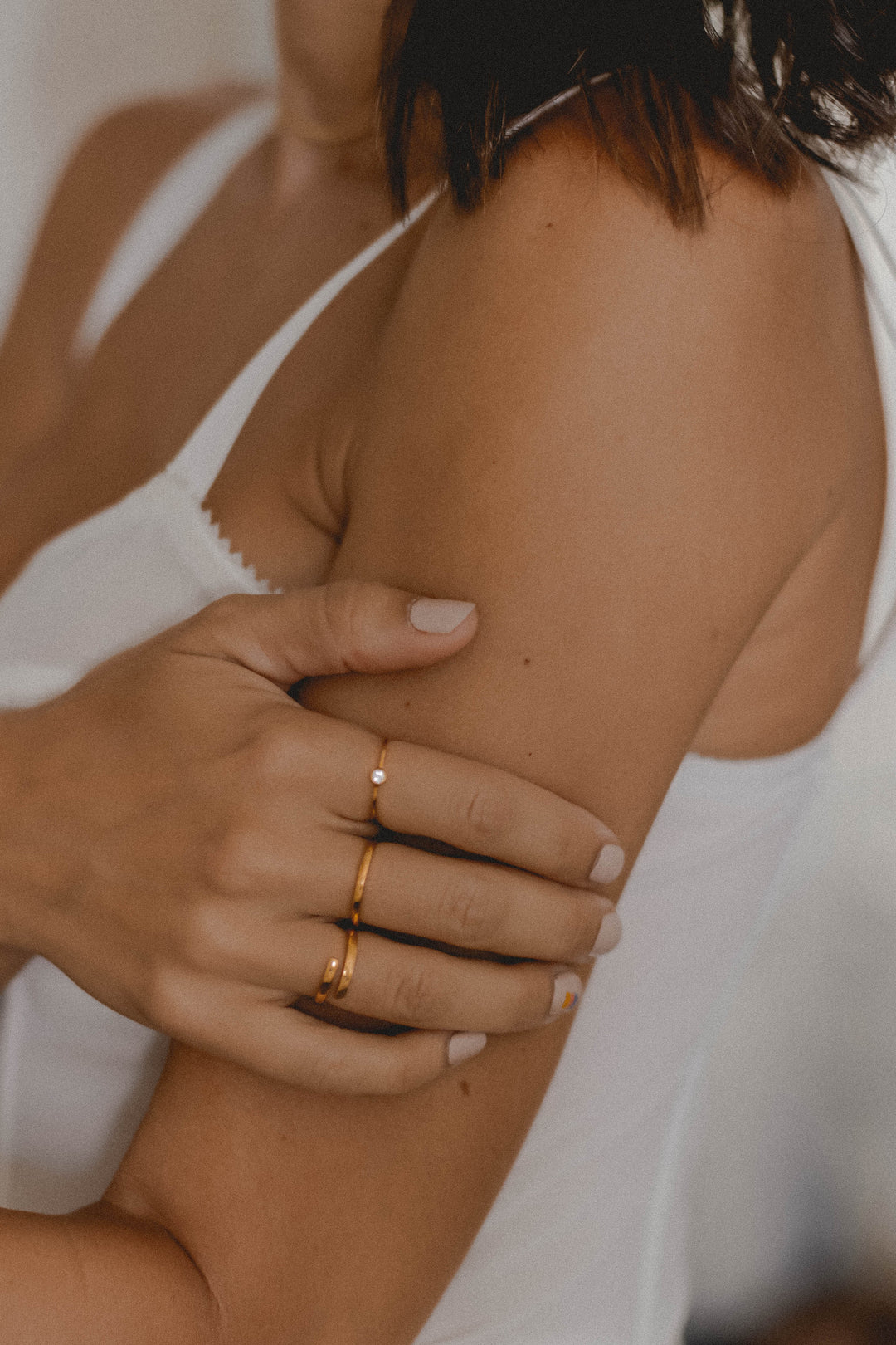 Close-up of a woman wearing delicate gold rings and a white tank top softly holding her upper arm against a neutral background