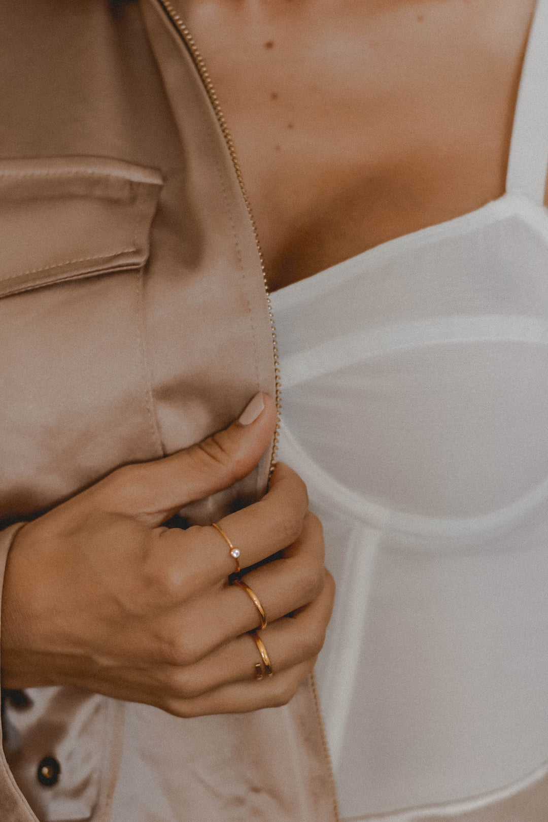 Close-up of a woman’s hand with gold rings holding a beige jacket open to reveal a white structured top