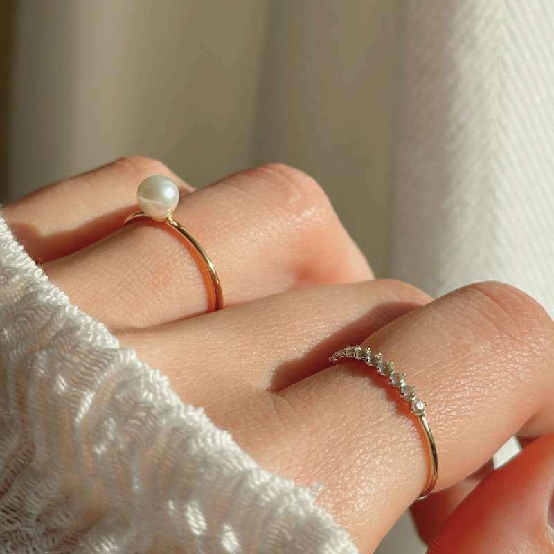 Close-up of a hand wearing two delicate gold rings, one with a small pearl and the other with tiny diamonds on a light background