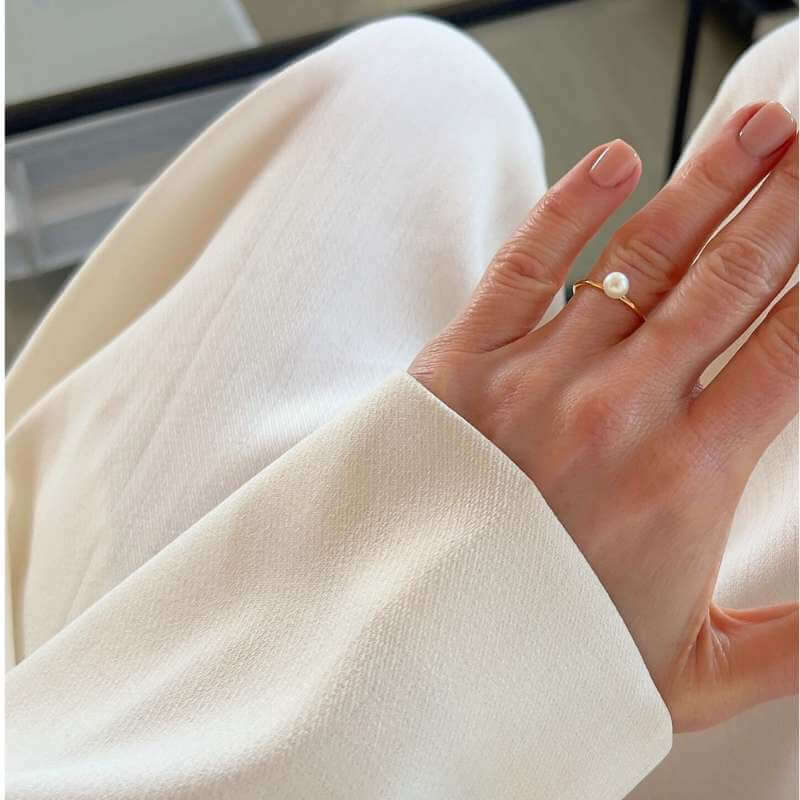 Close-up of a hand wearing a delicate gold ring with a single pearl against soft white fabric background