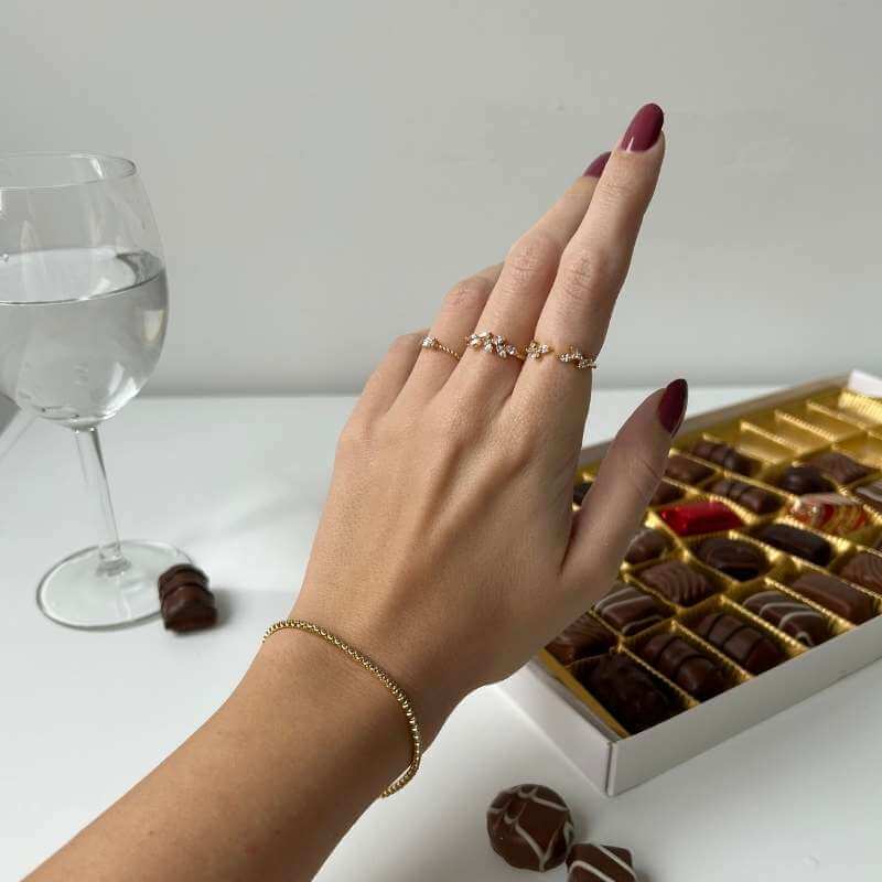 Close-up of a hand with dark red nails wearing delicate gold rings and bracelet next to chocolates and a glass of water