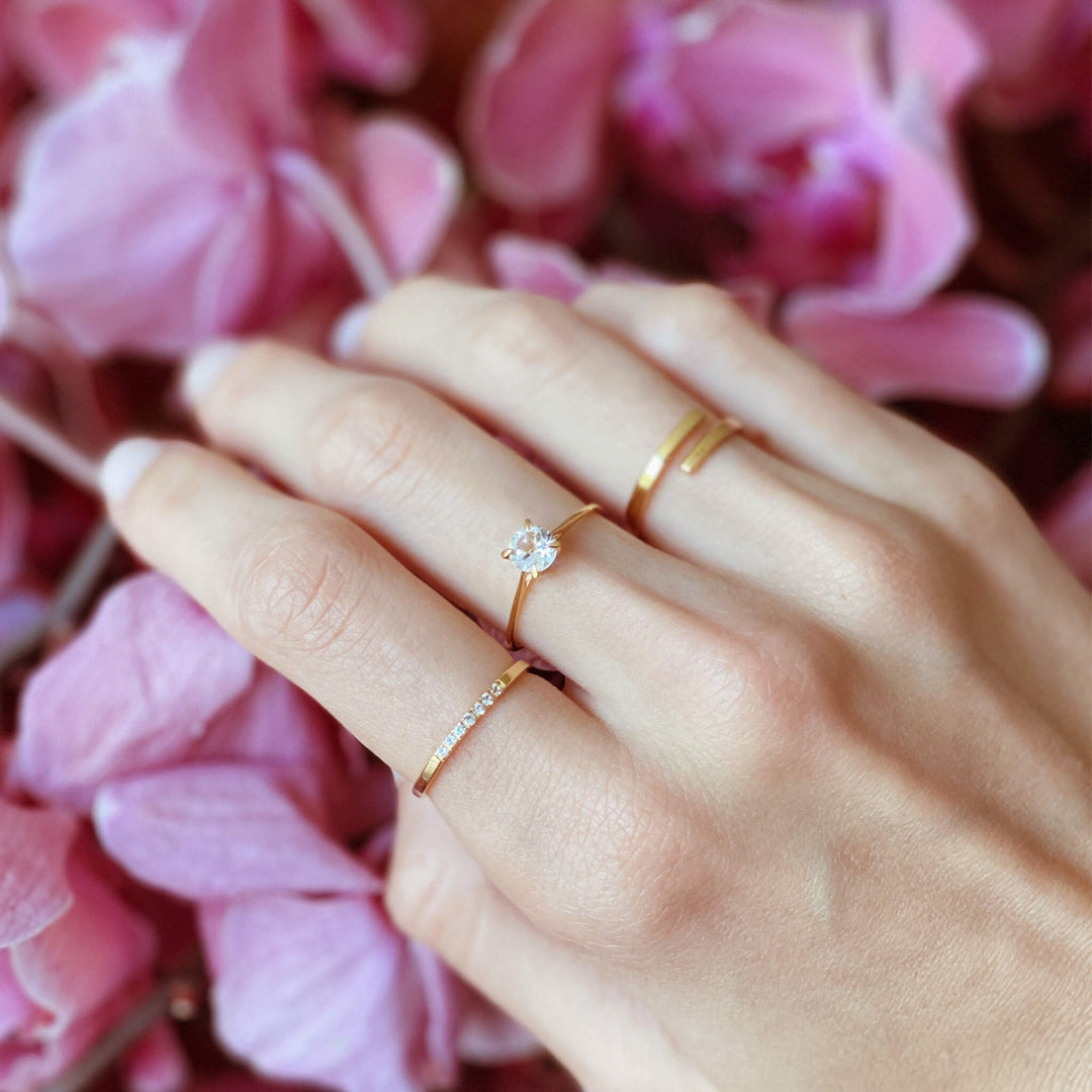 Close-up of a hand with three delicate gold rings, including one with a clear round stone, against pink flower petals background