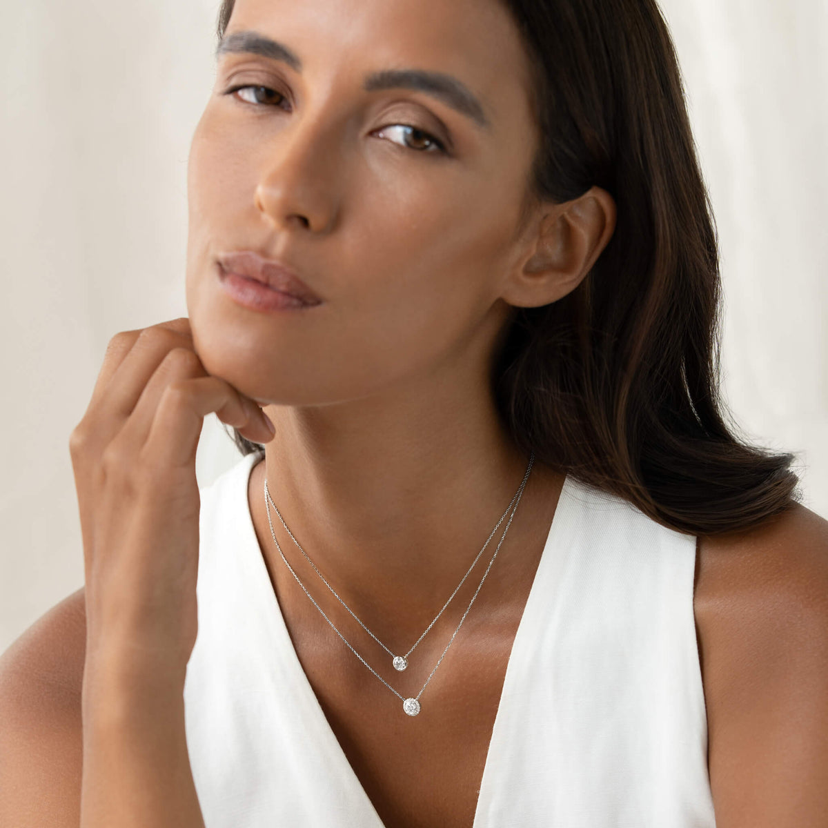 A woman in a white top rests her hand on her chin, wearing layered delicate white gold necklaces. The shorter necklace features a small diamond pendant in a bezel setting, while the longer one has a round brilliant-cut diamond halo pendant.