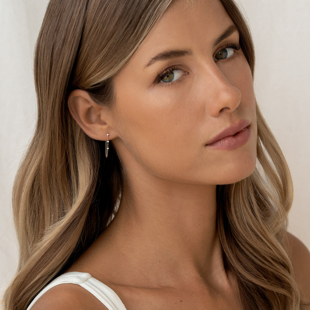A woman with long, wavy brown hair and a calm expression wears a small, Gold delicate earring. Her neutral-toned outfit complements the serene background.