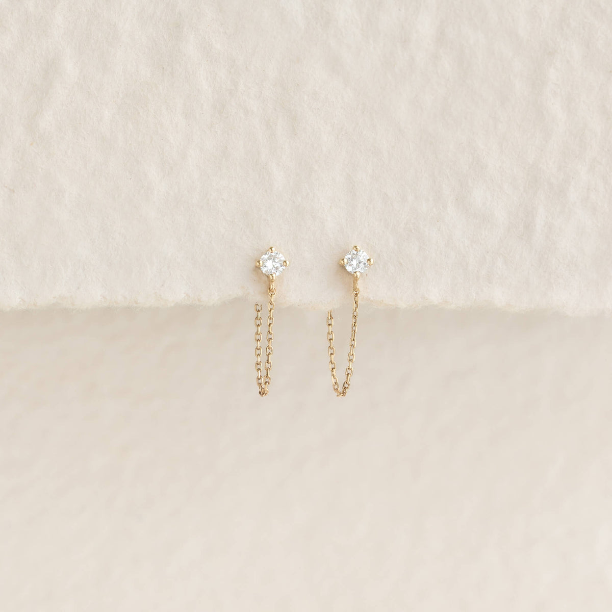 Delicate Gold earrings with sparkling stones and thin chains hang against a soft, textured beige background, conveying elegance and simplicity.