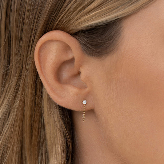 Close-up of a woman's ear wearing a Gold stud chain earring with a small diamond stud. The background is simple, highlighting the elegant jewelry.