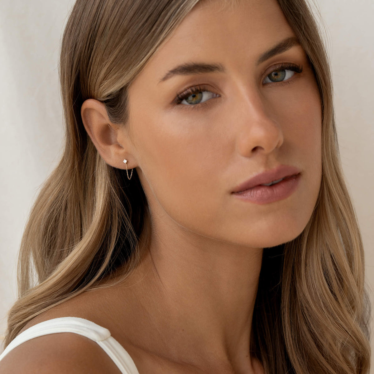 Woman with long light brown hair and soft expression wearing a white top and small Gold chain earring, set against a simple cream background.