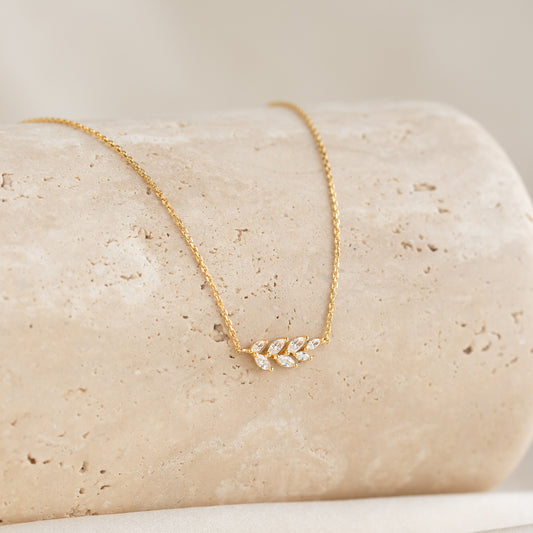 Gold necklace with a delicate leaf pendant adorned with small diamonds, elegantly displayed on a textured beige stone. The tone is elegant and refined.