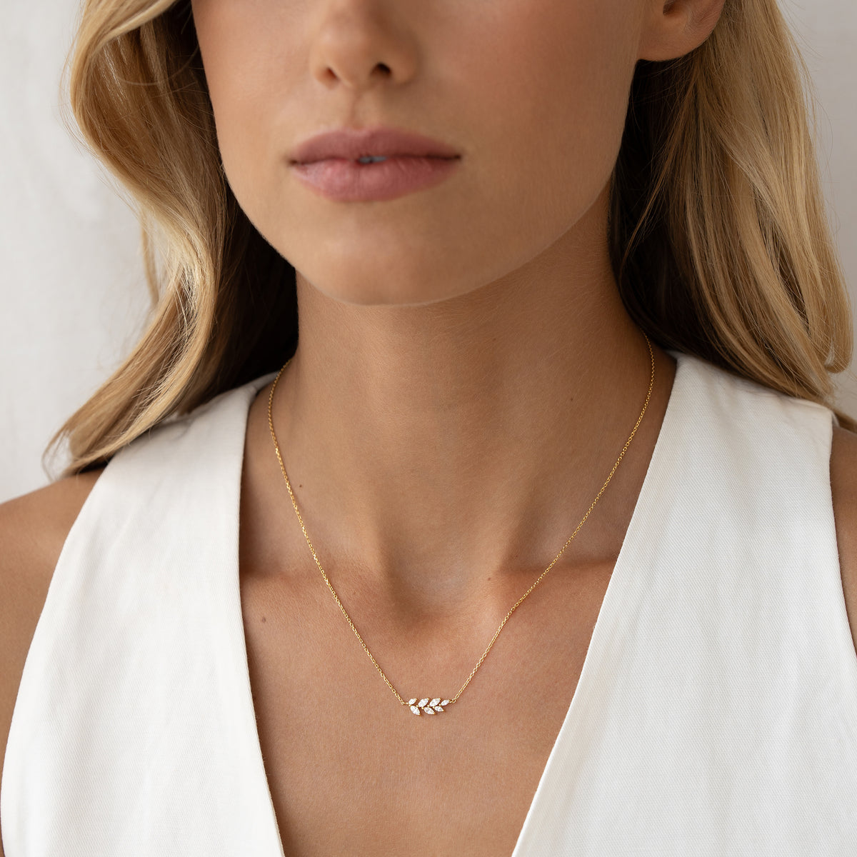 A woman wearing a delicate Gold necklace with a leaf-shaped pendant against a white sleeveless top. The tone is elegant and minimalist.