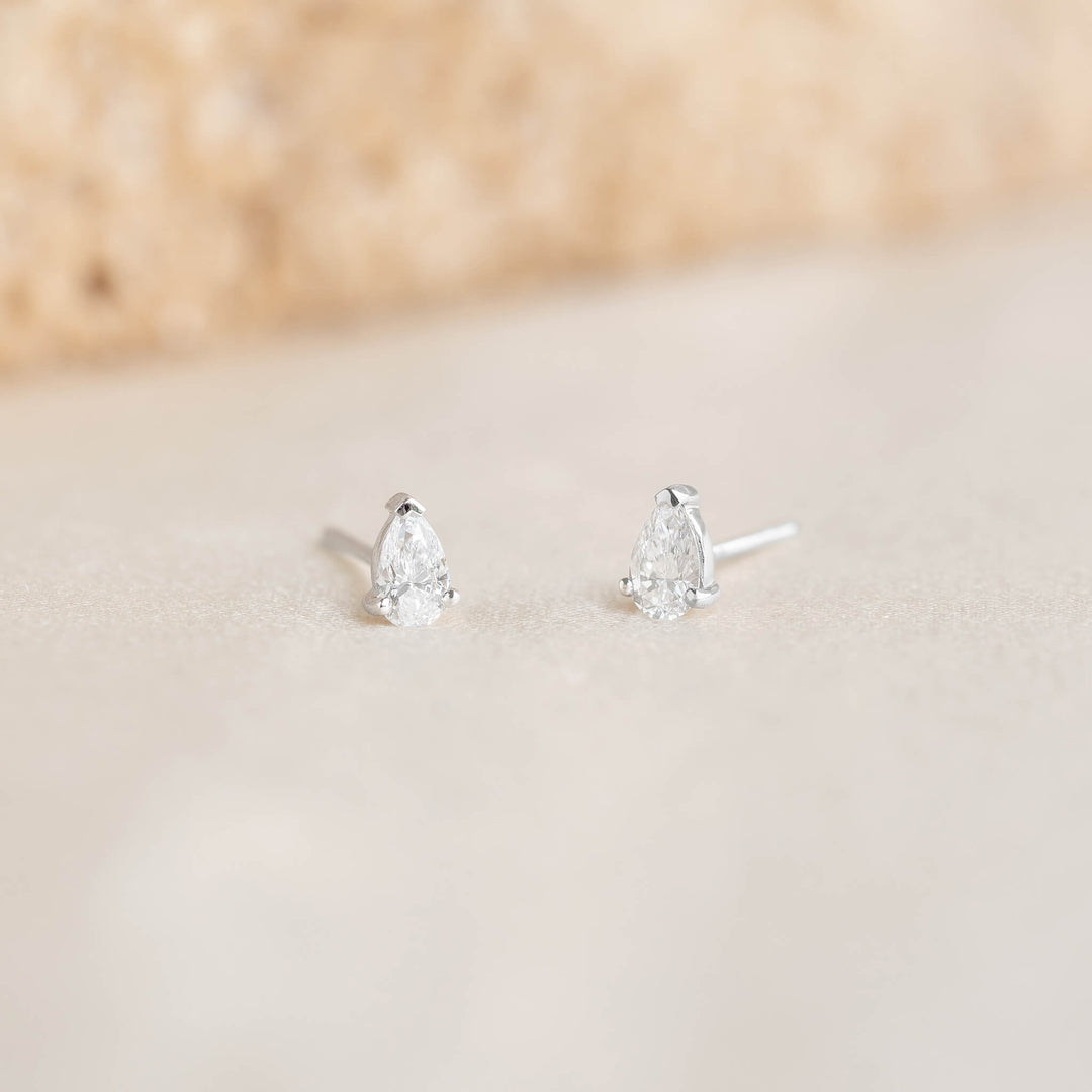 Two White Gold pear-shaped diamond stud earrings on a soft, blurred background. The earrings' simplicity conveys elegance and subtle sophistication.