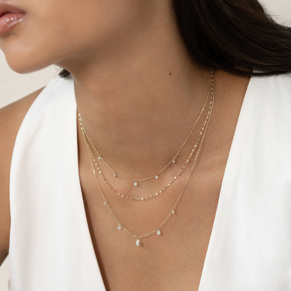 A person wearing three layered Gold necklaces with small dangling pendants, paired with a sleeveless white outfit, giving a minimalist and elegant look.