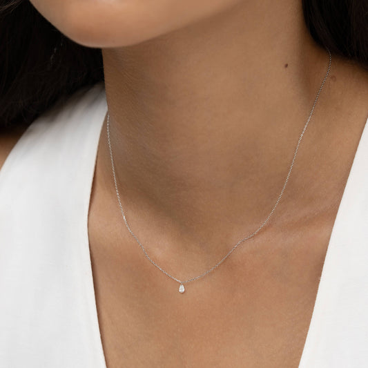 A delicate White Gold necklace with a small teardrop pendant adorns a woman's neck. She wears a white top, and the scene conveys elegance and simplicity.