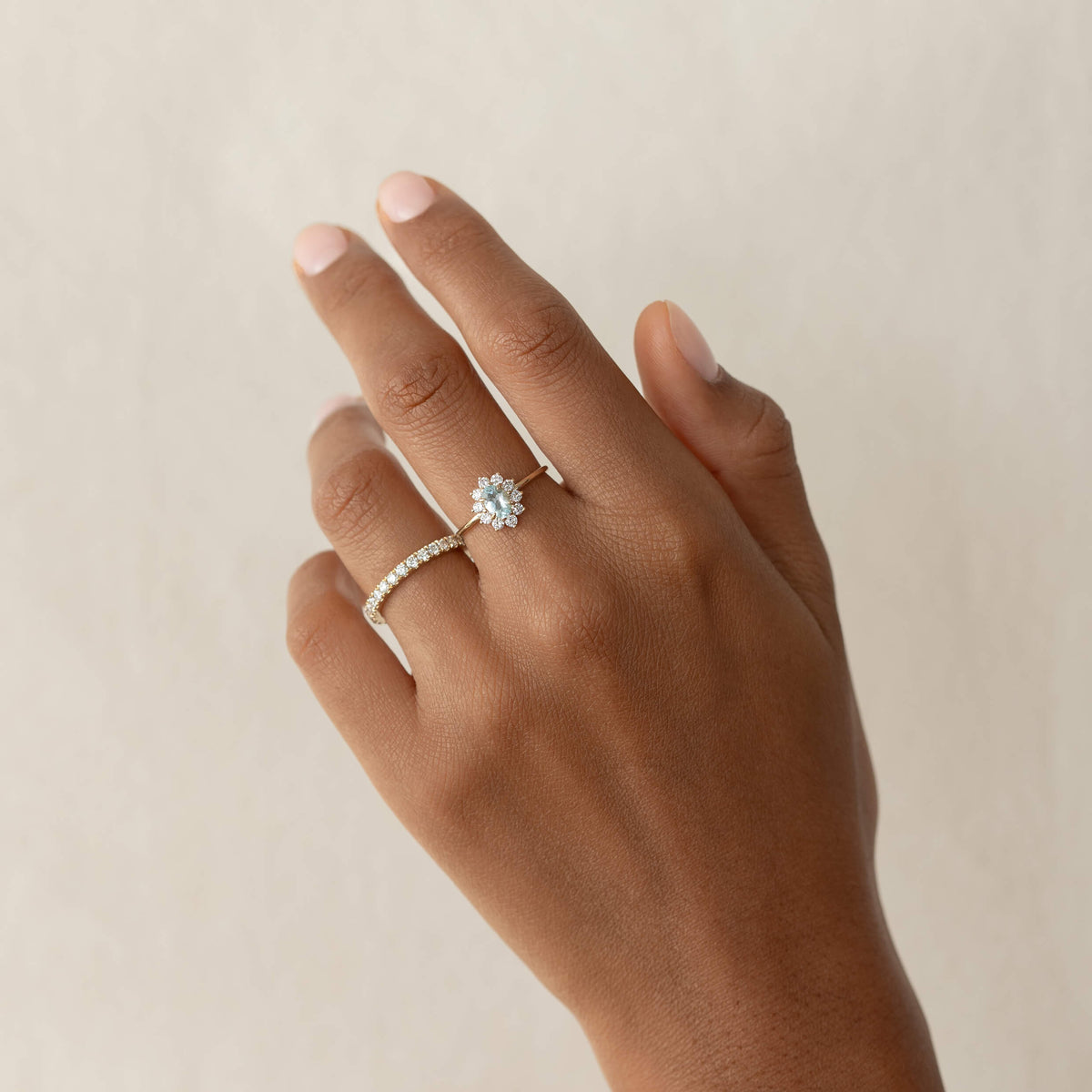A hand with a soft beige background displays Gold two rings; a diamond floral design on the ring finger and a simple, sparkling band on the middle finger.