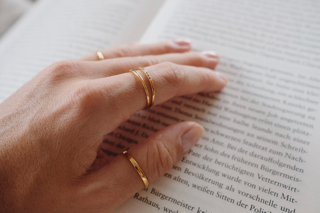 Close-up of a hand with minimalist gold rings resting on an open book with blurred text on a white background