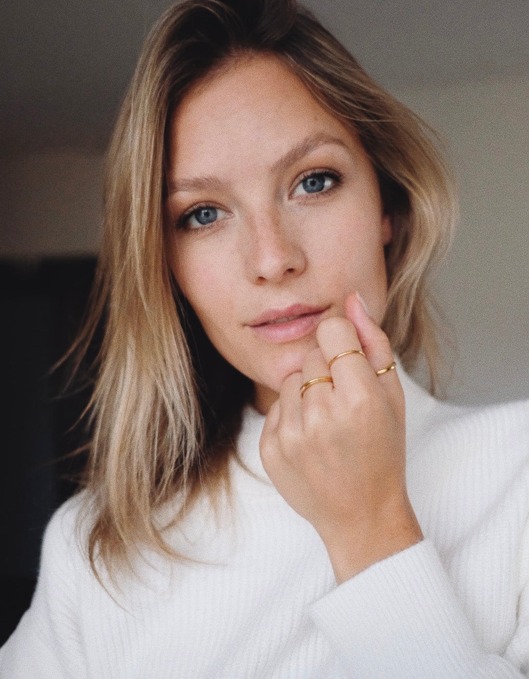 Close-up of a woman with light blonde hair wearing a white ribbed top and minimal gold rings on her fingers