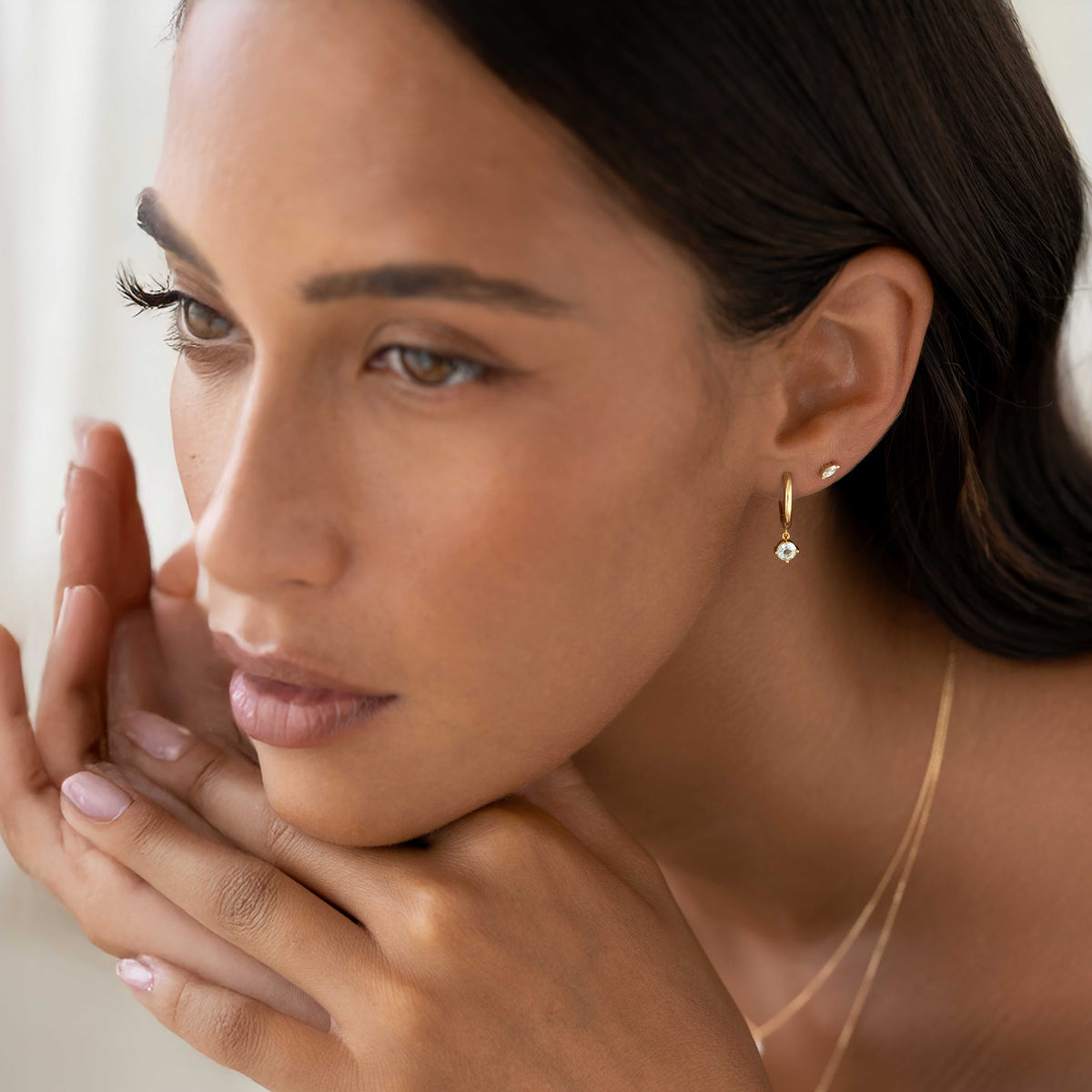 Close-up of a woman wearing March Birthstone Drop Huggie Earrings - Aquamarine with delicate gold chains on a neutral background