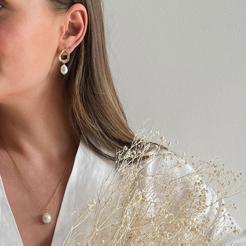 Close-up of a woman wearing a gold earring with a teardrop pearl and a matching pearl pendant necklace against a white background