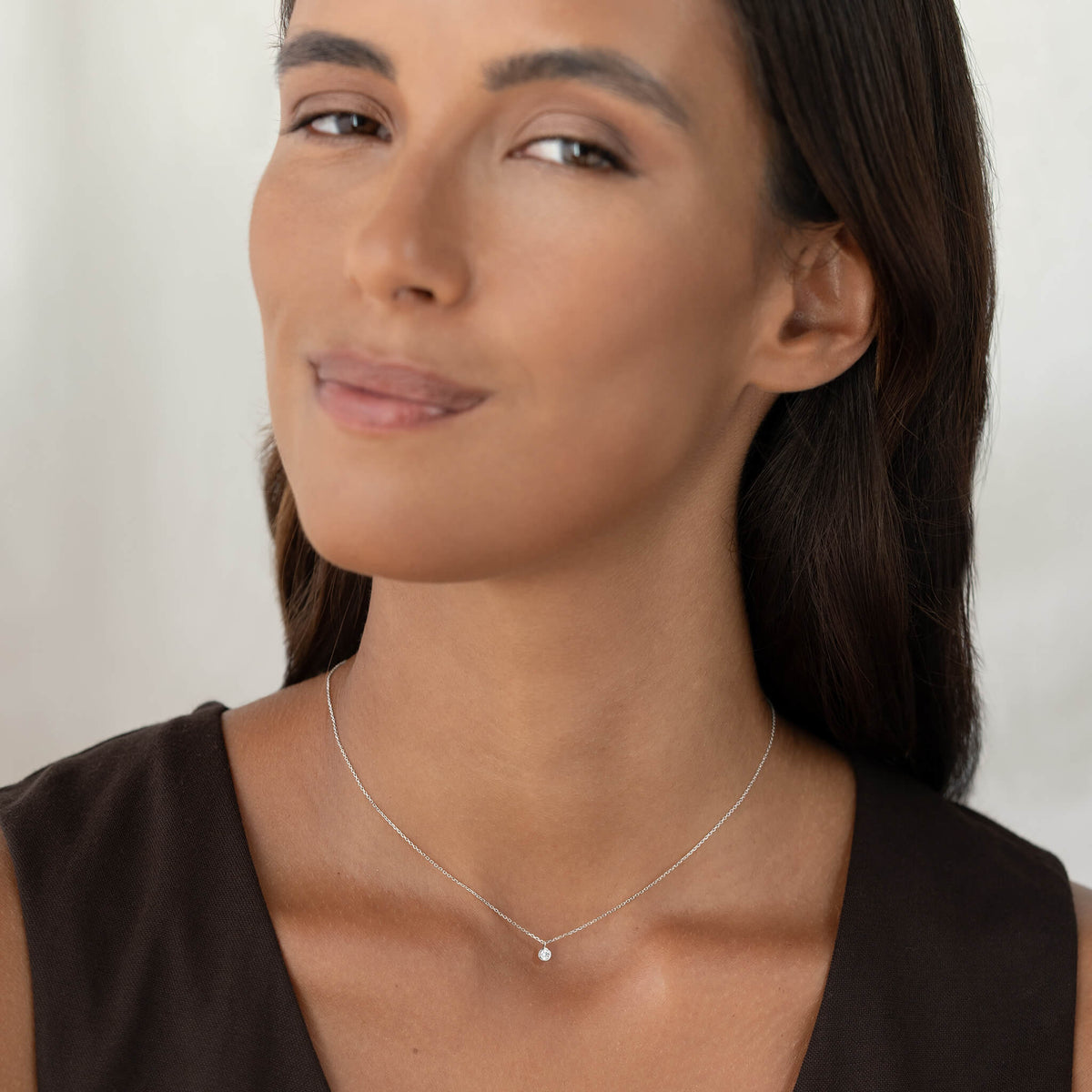 A close-up portrait of a woman with dark hair wearing a minimalist, delicate White Gold chain necklace with a tiny round brilliant diamond pendant, styled with a simple dark brown top.