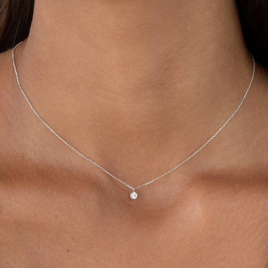 A close-up shot of a person wearing a very delicate White Gold necklace with a tiny, round brilliant diamond solitaire pendant set in a minimalist bezel setting.