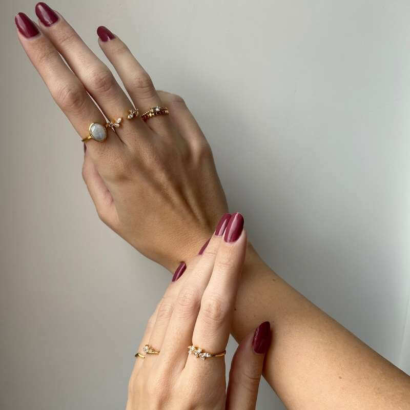 Close-up of two hands with glossy dark red nail polish wearing delicate gold rings with small gemstones on a neutral background