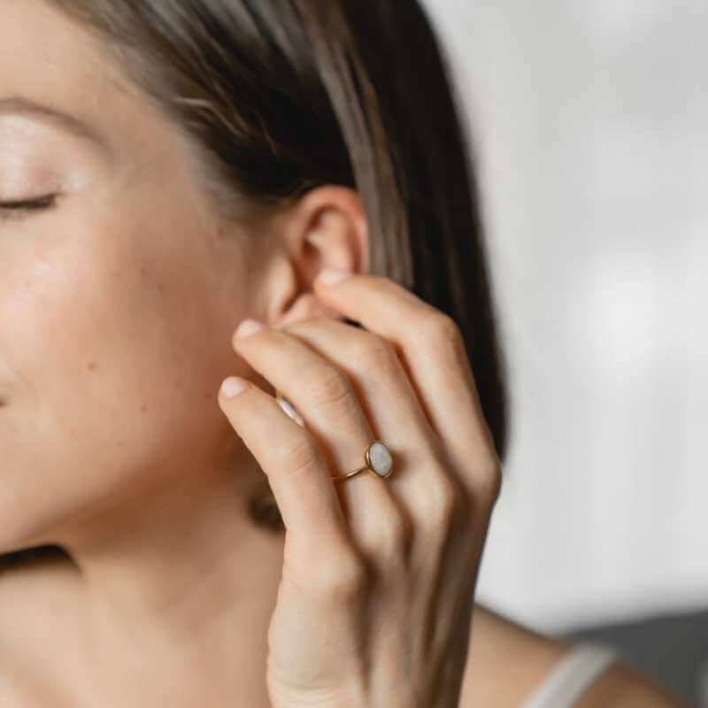Close-up of a woman with closed eyes touching her ear, wearing a delicate gold ring with a white oval stone