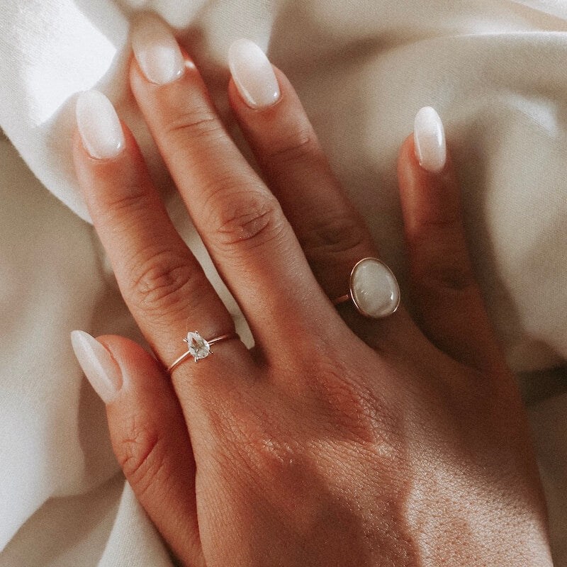 Close-up of a hand with almond-shaped nude nails wearing delicate silver rings on a soft white fabric background