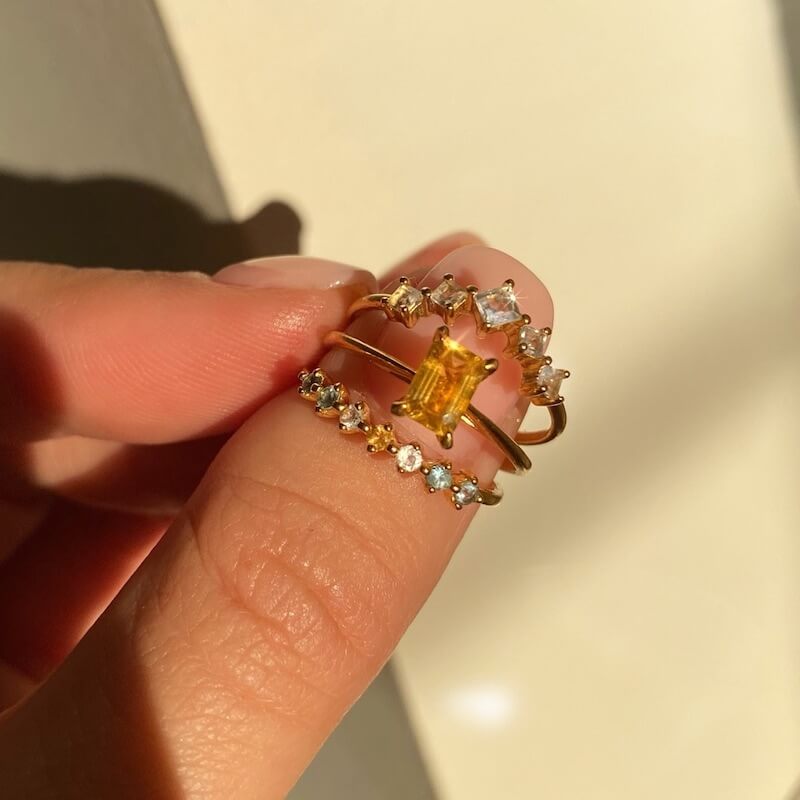 Close-up of a hand holding three gold rings with white and yellow gemstones against a soft beige background