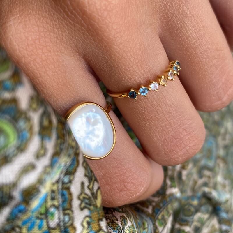 Close-up of a hand wearing a gold ring with a large white stone and a delicate gold ring with multiple small blue and yellow gems