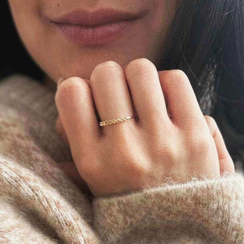 Close-up of a hand wearing a delicate gold ring with small round yellow stones against a soft beige sweater background