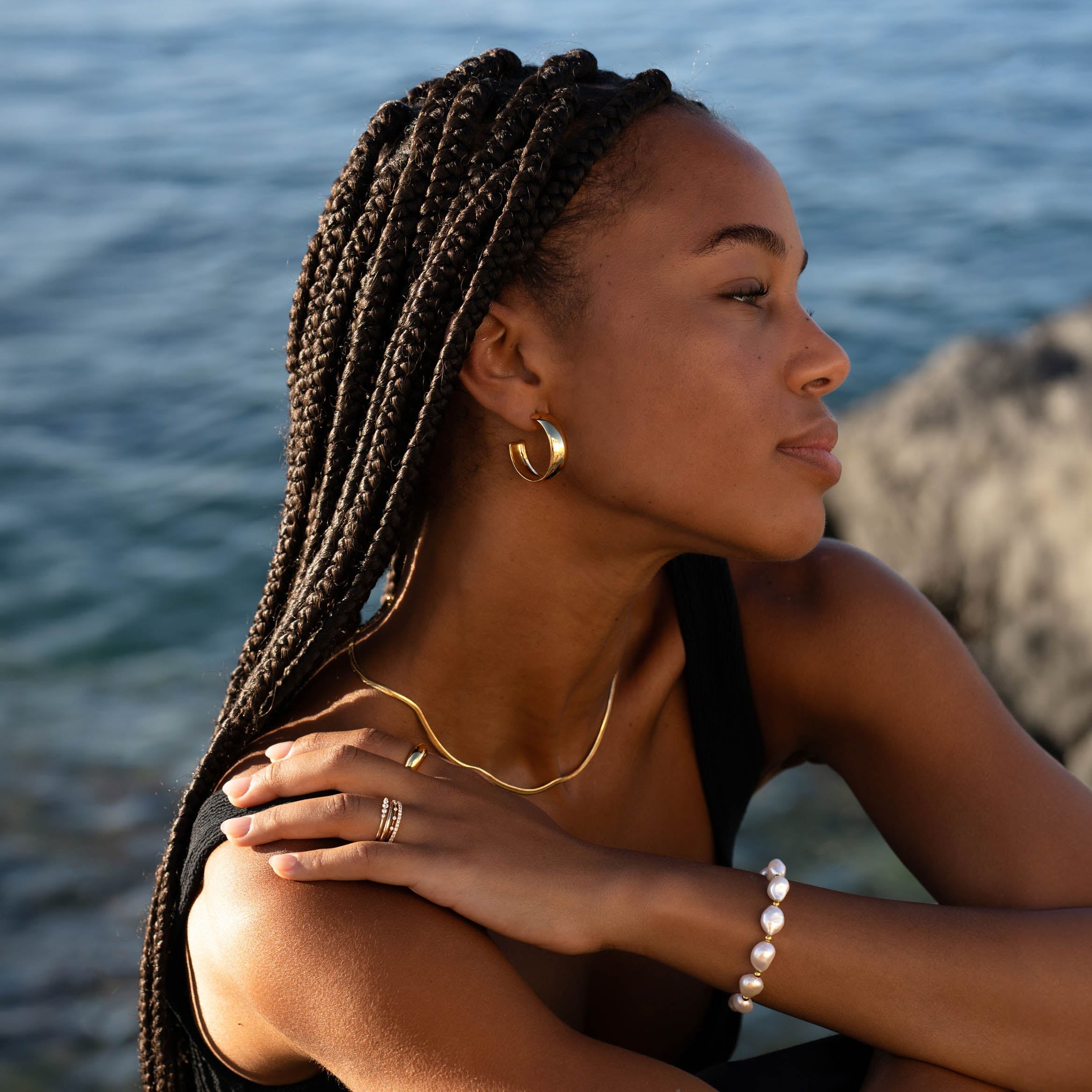 Woman wearing Large Bold Hoop Earrings - Mina with gold necklace and pearl bracelet by the water in natural light