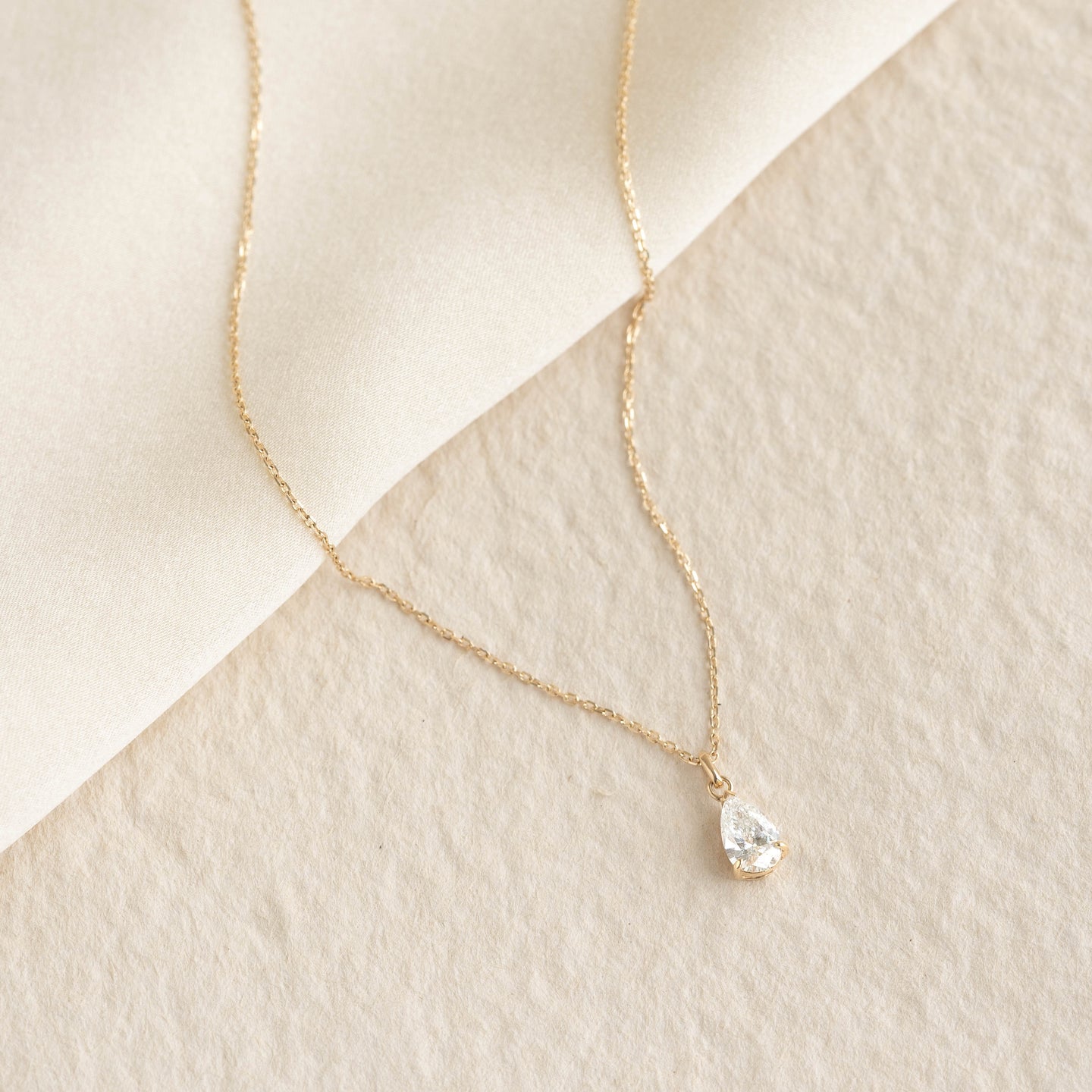 Pear Cut Diamond Pendant Necklace 14k Gold - Oline with a delicate chain on a soft beige textured background