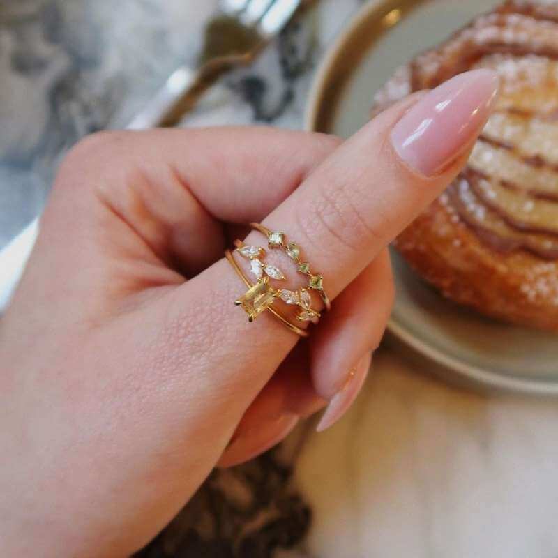 Close-up of a hand with light pink nails wearing three delicate gold rings with colored gemstones over a marble surface background