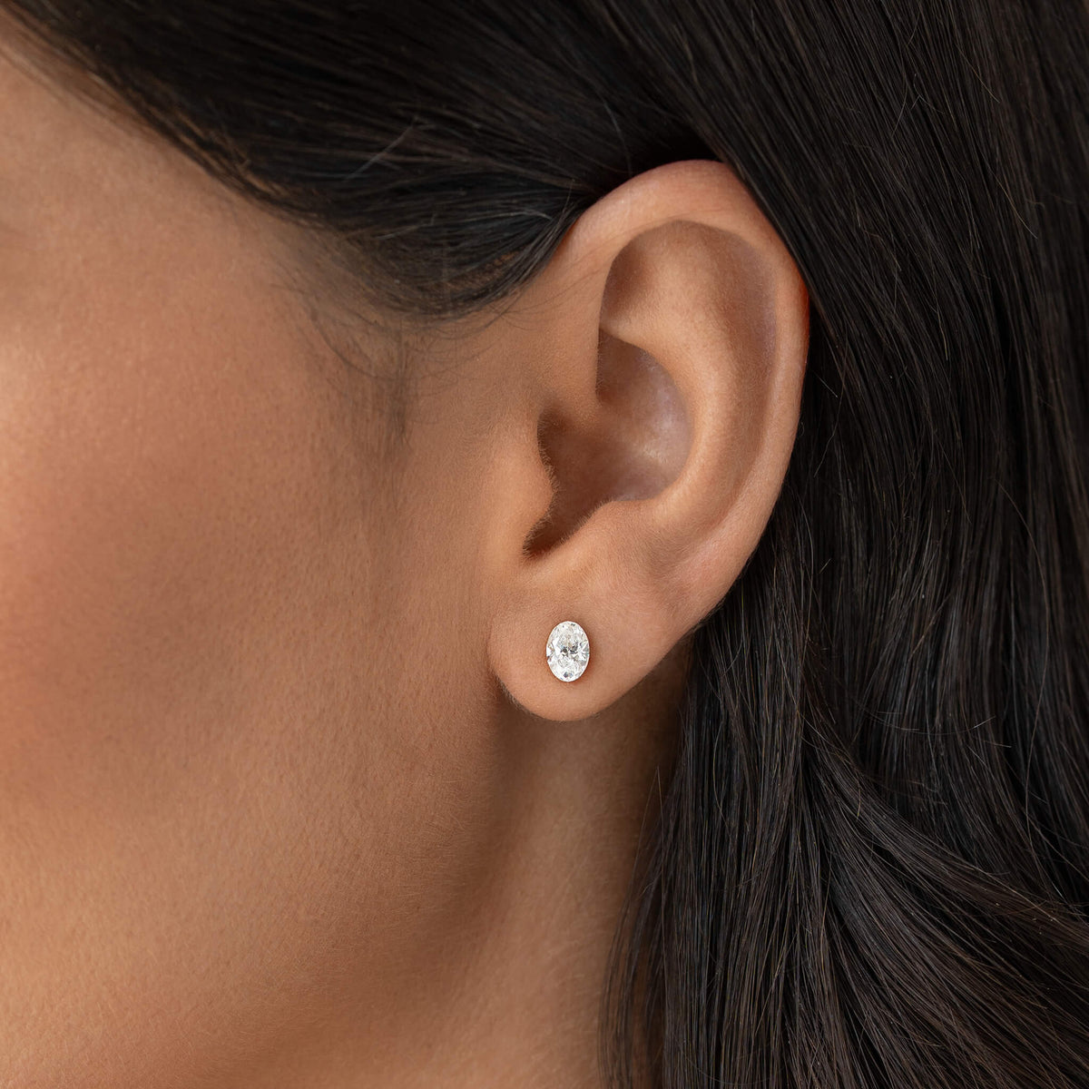 Close-up of a Woman's ear wearing a small Gold oval diamond stud earring set in Gold metal, with smooth skin and dark hair tucked behind the ear.