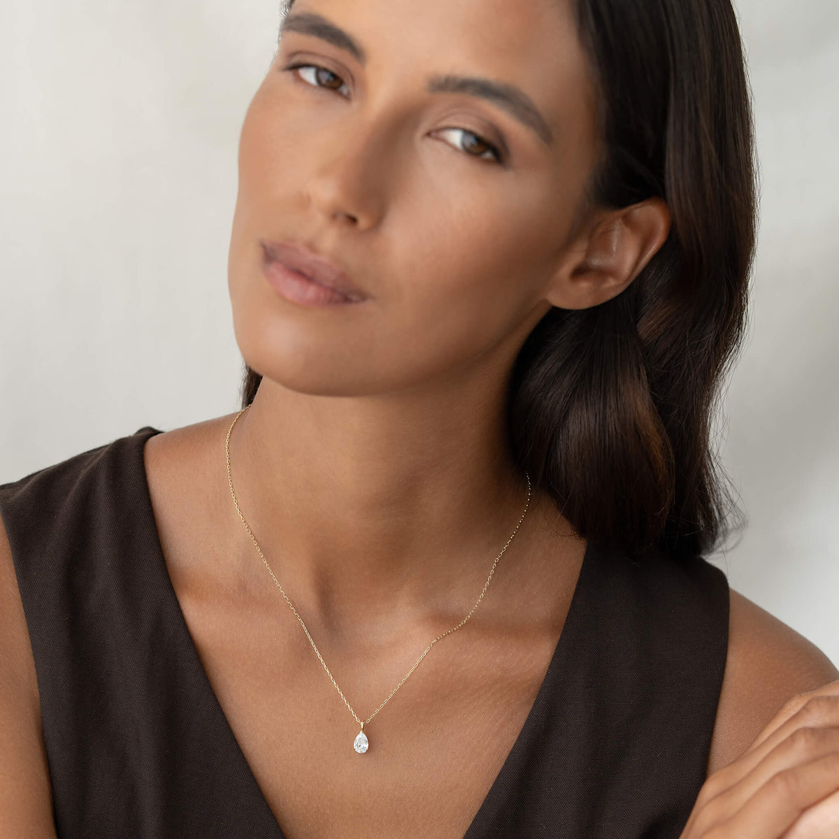 A portrait of a woman with dark hair wearing a minimalist Gold necklace with a single pear-shaped diamond pendant, set against a simple, elegant dark top.