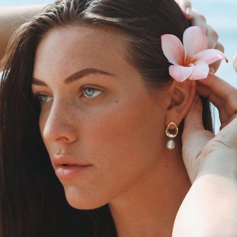Close-up of a woman with light freckles wearing a pearl drop earring and holding a pink flower near her hair by the ocean