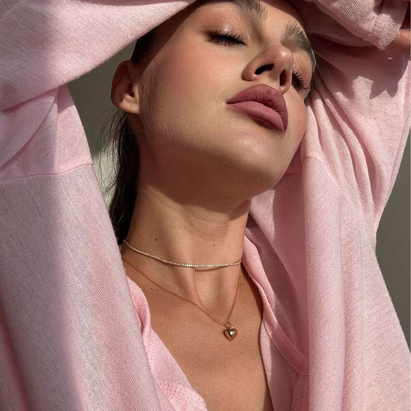 Close-up of a woman with closed eyes wearing layered delicate necklaces and a soft pink top against a neutral background