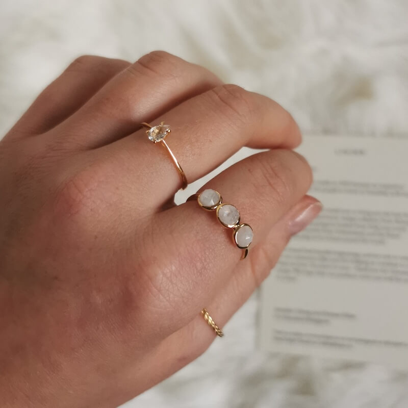 Close-up of a hand wearing three delicate gold rings with small gemstones on a soft white background