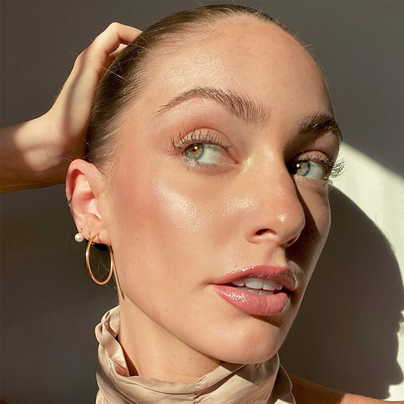 Close-up of a woman with pulled-back hair wearing a gold hoop earring and small pearl stud, with natural light shadows