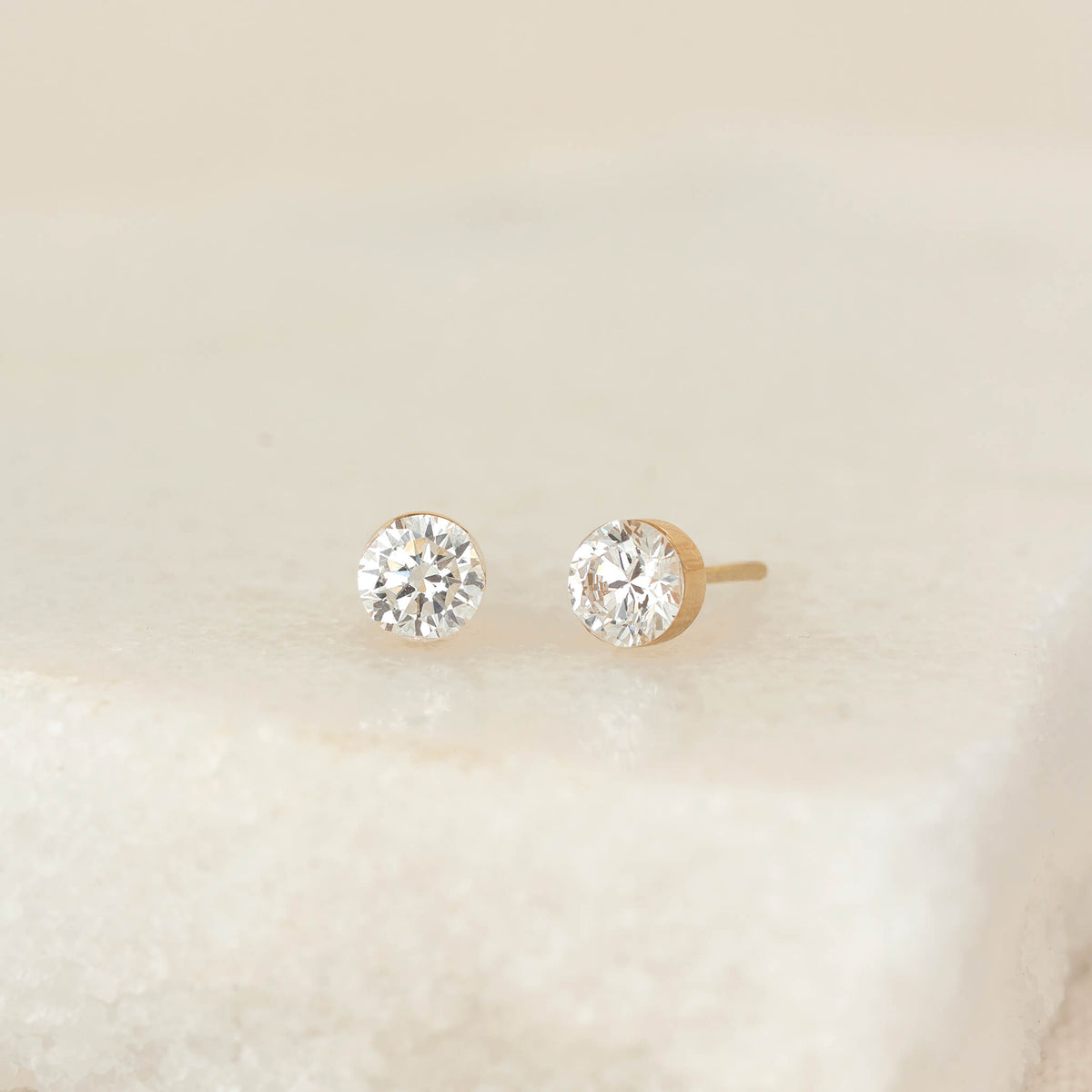 Pair of round Gold diamond 1 ct tw stud earrings on a neutral surface, featuring brilliant-cut stones in simple settings with one earring showing the post.