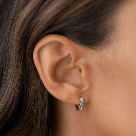 Close-up of an ear wearing September Birthstone Huggie Earrings - London Blue Topaz with gold vermeil setting on a neutral background