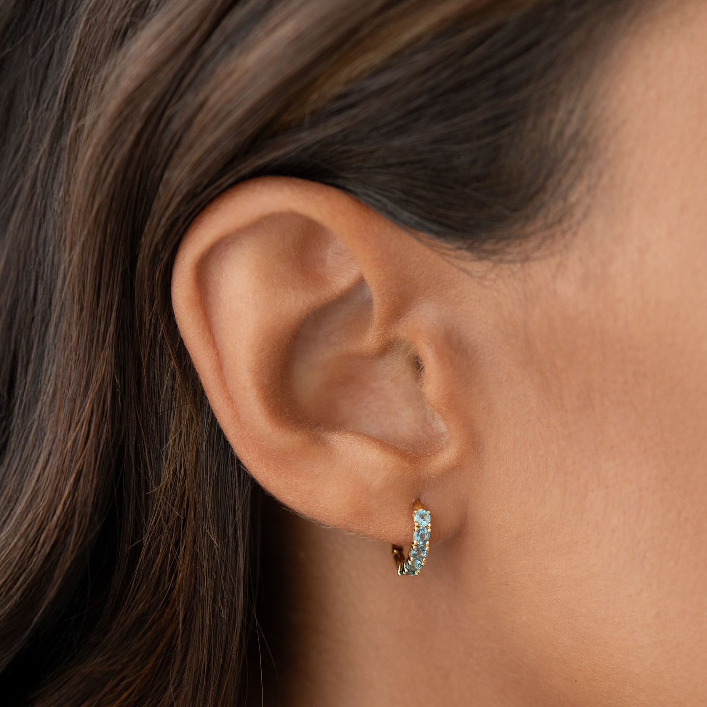 Close-up of an ear wearing September Birthstone Huggie Earrings - London Blue Topaz with gold vermeil setting on a neutral background
