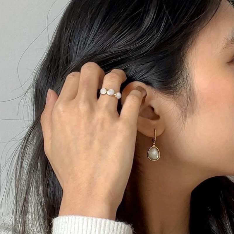 Close-up of a woman with dark hair wearing a pearl ring and a teardrop-shaped pearl earring against a light gray background