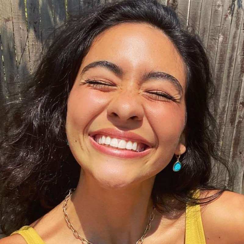 Close-up of a woman with curly dark hair smiling with eyes closed wearing a turquoise drop earring and yellow top outdoors