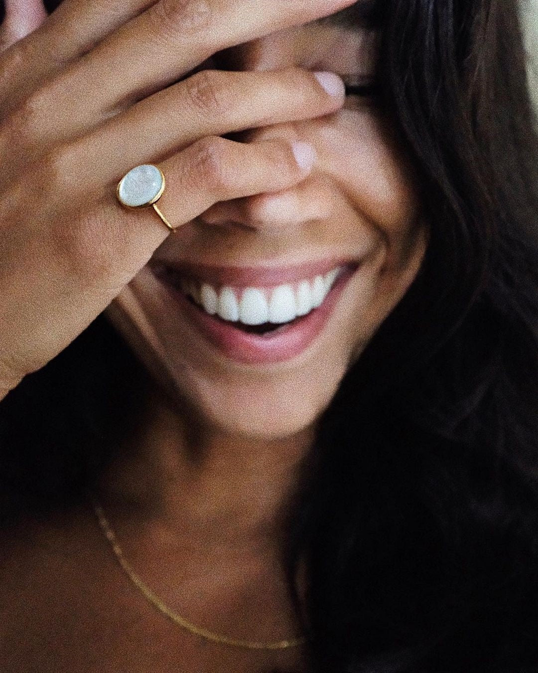 Close-up of a smiling woman partially covering her face with her hand, wearing a gold ring with a white stone and a delicate gold necklace