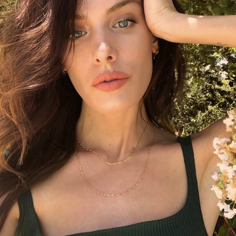 Close-up of a woman with loose brown hair wearing coral lipstick and layered gold necklaces outdoors in natural sunlight