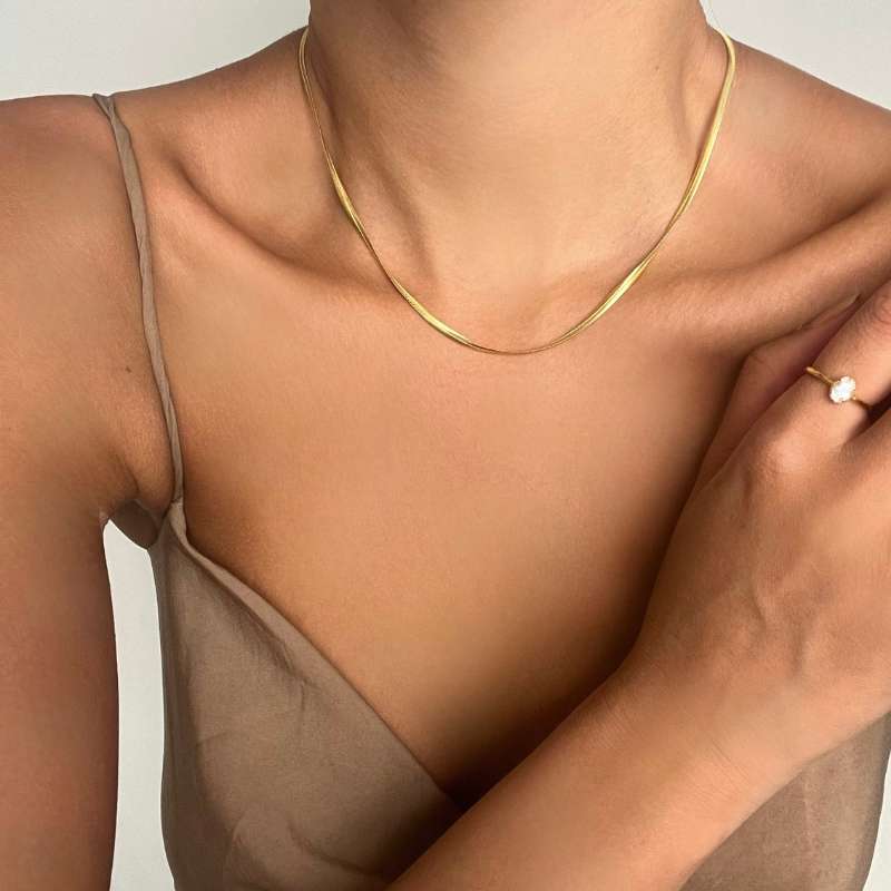 Close-up of a woman wearing a thin gold chain necklace and a gold ring with a white stone on her hand against a neutral background